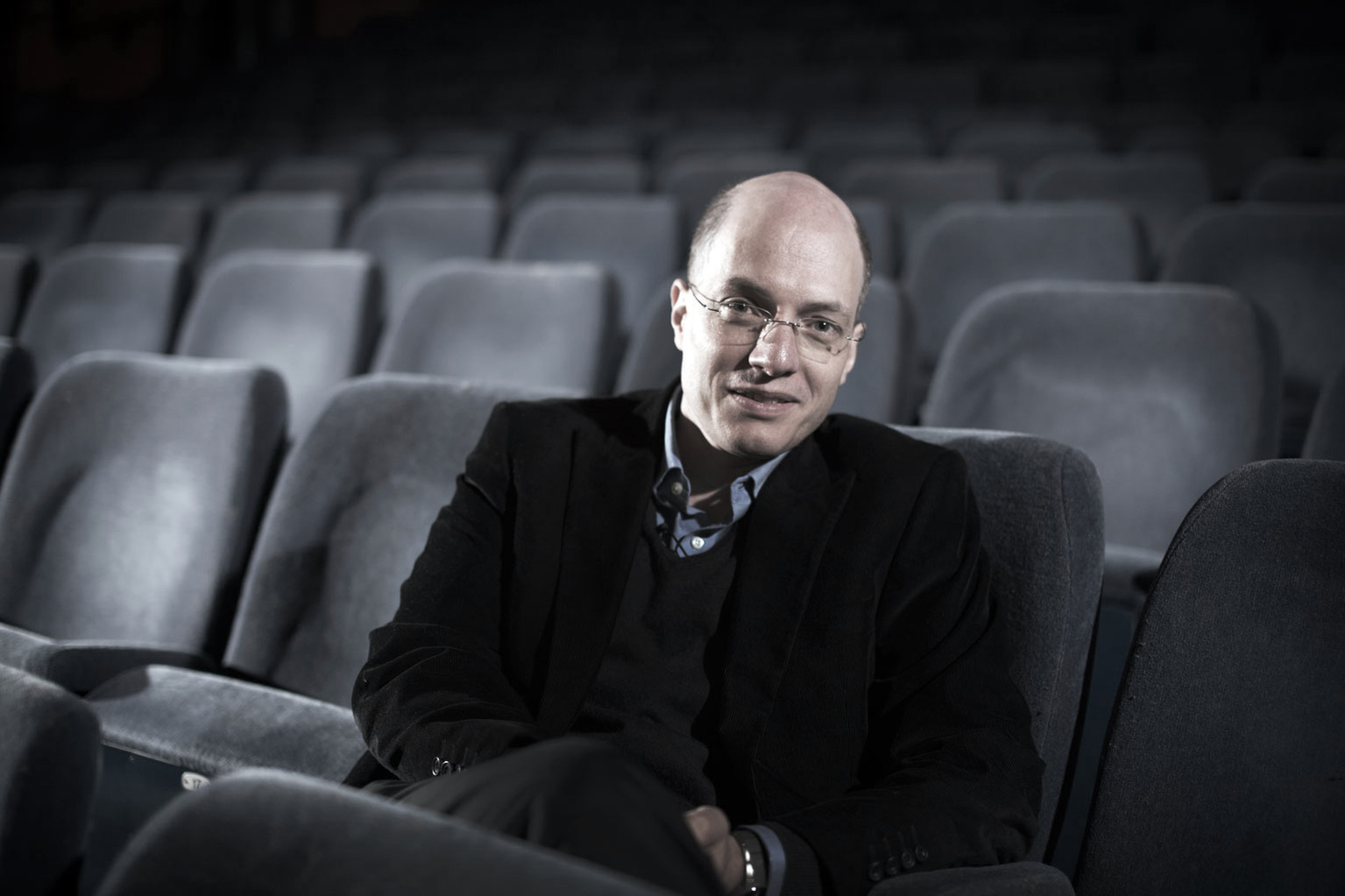 Portrait of the British-Swiss philosopher Alain de Botton in a theatre alone. The theatre seats are dark grey and he is wearing a black suit. Shot by Norwich commercial, freelance portrait photographer Matt Keal.