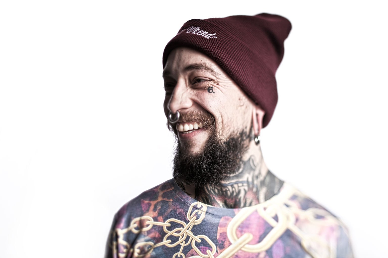 Studio portrait of a man with tattoos, a beard and a nose ring. He's wearing a hat and smiling to the left. Shot by Norwich commercial, freelance portrait photographer Matt Keal.