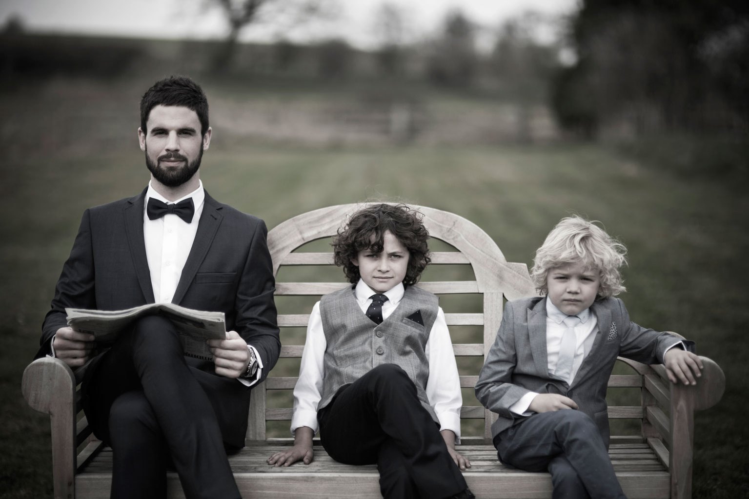 A bearded man and two children dressed in suits are sitting on a wooden bench outside facing the camera for a fashion campaign photoshoot. Shot by Norwich commercial, freelance portrait photographer Matt Keal.