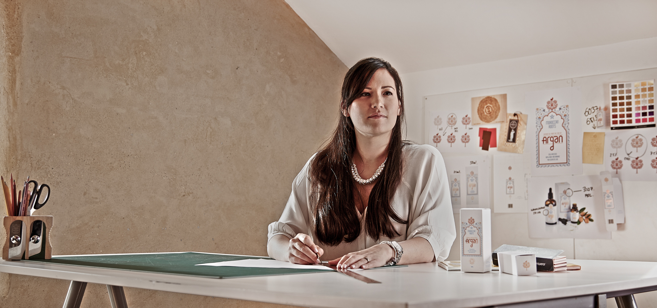 A young, female graphic designer working at her desk in a design studio. She is looking over the cameraman's shoulder and gazing into the distance. Shot by Norwich commercial, freelance portrait photographer Matt Keal.