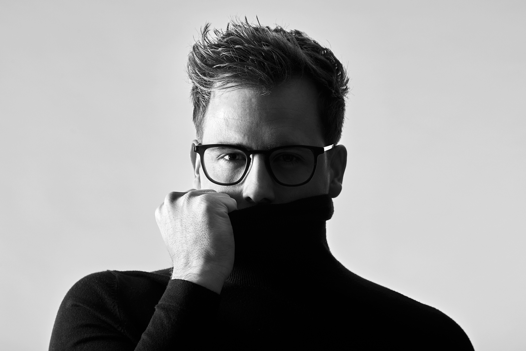 Headshot of a man pulling his roll neck jumper up to cover his mouth. He has short hair and is wearing thick-framed glasses. Shot by Norwich commercial, freelance portrait photographer Matt Keal.