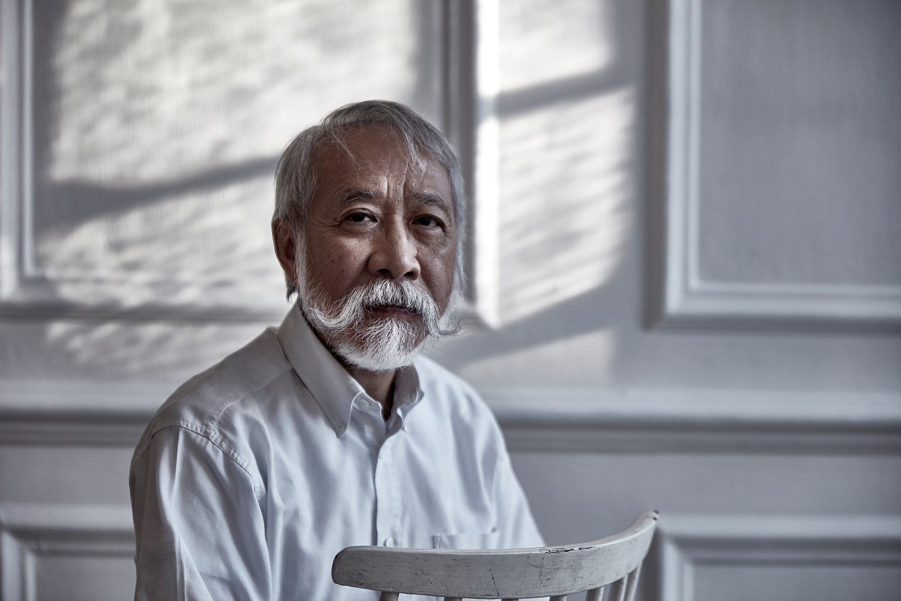 An old man with a grey handlebar moustache and beard wearing a white shirt and sitting on a painted white chair against a white panelled wall. Shot by Norwich commercial, freelance portrait photographer Matt Keal.