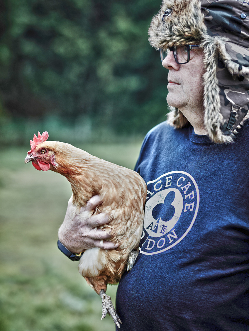 Informal portrait of a man with a fur-lined hat standing outside holding a chicken in his garden. Shot by Norwich commercial, freelance portrait photographer Matt Keal.