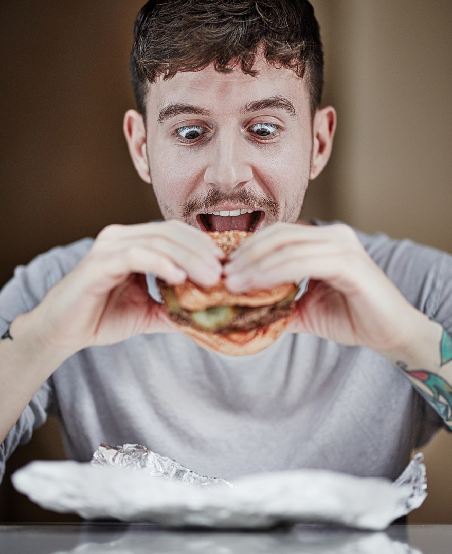 Portrait of a young man about to take a bite of a burger with tin foil on the table below. Shot by Norwich commercial, freelance portrait photographer Matt Keal.