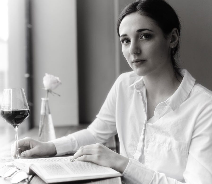 Corporate headshot of a restaurant owner or head chef, reading a book at a table with a glass of wine. Her hair is pulled back into a ponytail & she's wearing a white shirt at the photoshoot location. Shot by Norwich commercial portrait photographer