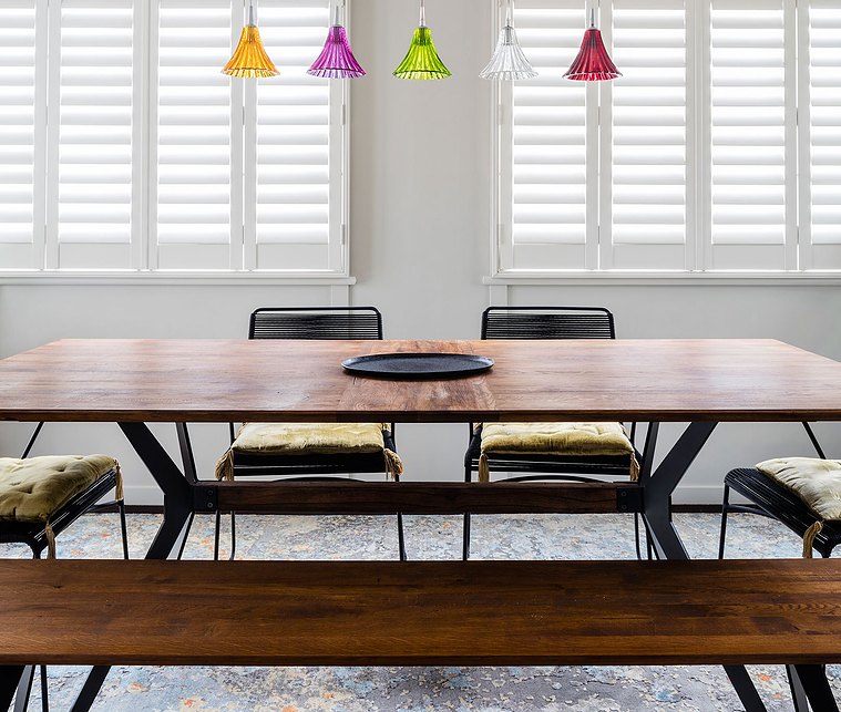 Colourful lamps hanging over a dark, wooden dining room table and black chairs with white shutters in the background