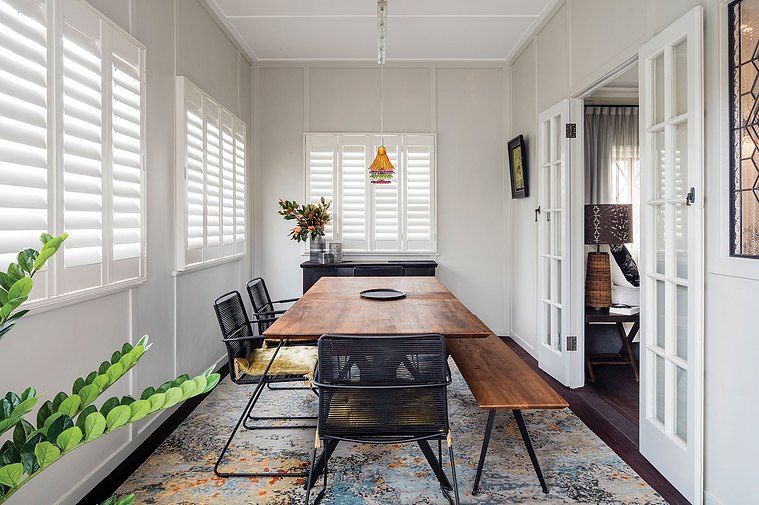 Colourful lamps hanging over a dark, wooden dining room table and black chairs with white shutters in the background