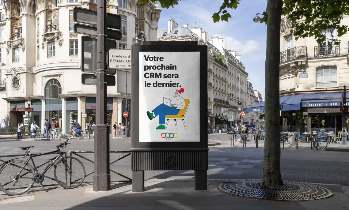 Affiche publicitaire, campagne Zoho : un personnage illustré, confortablement installé dans un fauteuil, se détend sur fond d'une rue parisienne baignée de soleil, combinant ainsi une ambiance urbaine et créative qui reflète l'esprit innovant de la marque