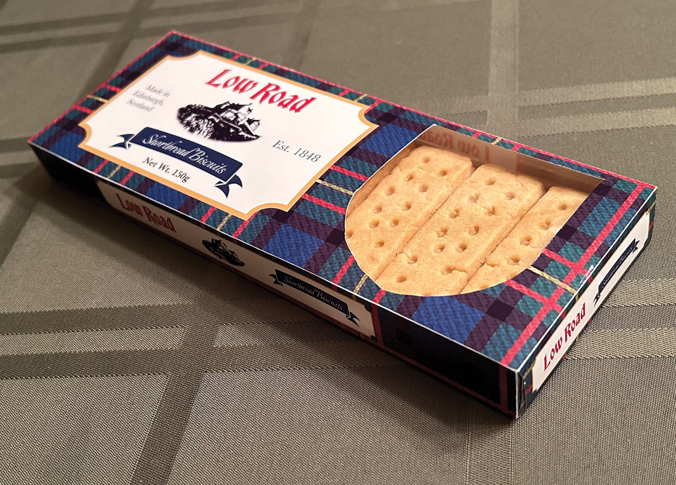 Low Road shortbread box with a shield shaped die cut revealing the biscuits inside sitting on tablecloth. 