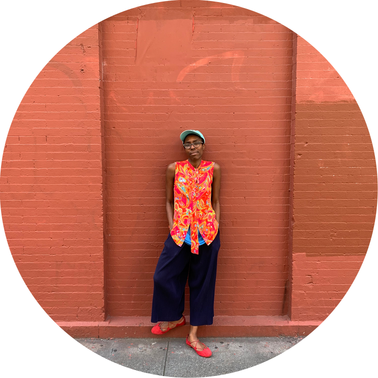 A Black woman wearing a baby blue baseball cap, dark glasses, a sleeveless paisley orange blouse, navy blue palazzo pants, and red flats stands casually against a red brick wall. 