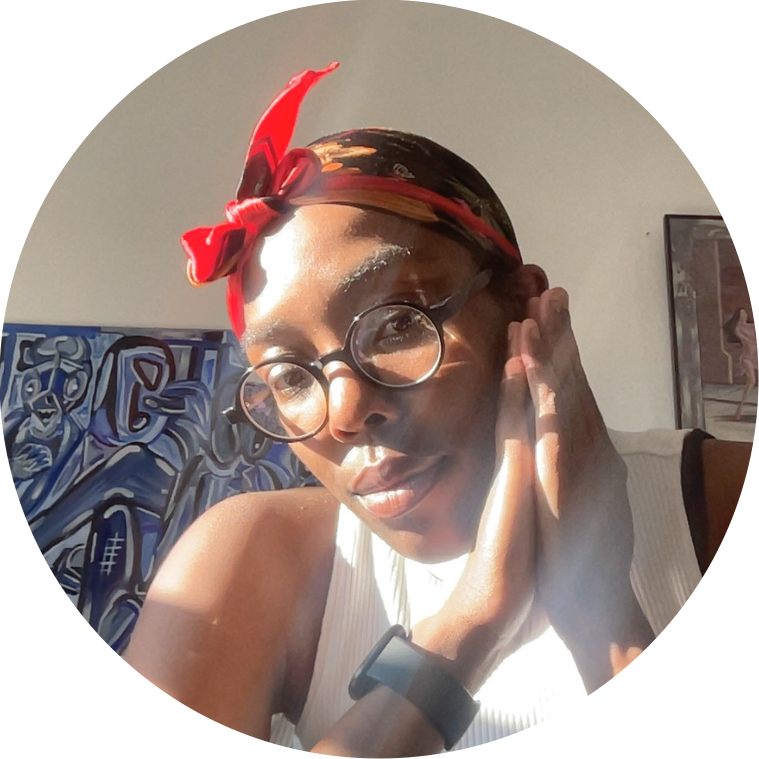 Dark skinned black woman sits  with her palms pressed together and resting on her cheek. On her left arm, a black smart watch. The woman wears a floral, black and red silk headscarf, black circle frame glasses, and a white shirt. Artworks hang behind her.