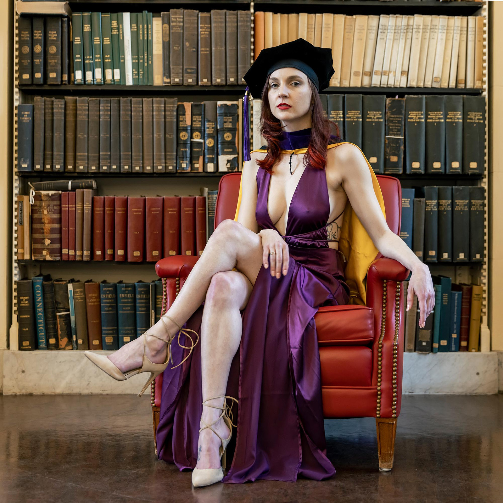 A woman sitting in an evening dress and graduate regalia in a library