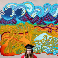 A BU graduate wearing cup and gown in front of mural in Boston.