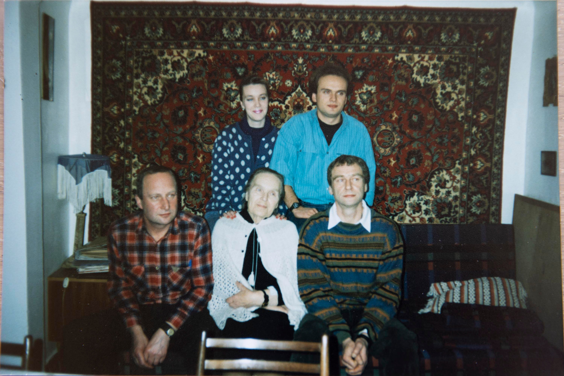 A vintage family photo shot on film in early 1990s. Group of five people, three are sitting on a couch and two are standing behind them in front of a carpet on a wall