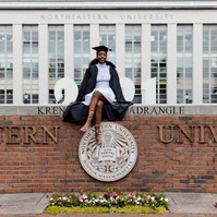 A Northeastern University graduate wearing cup and gown sitting on a wall in Boston.
