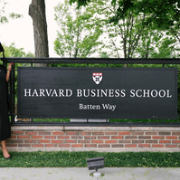 A graduate wearing cup and gown in front of Harvard Business School sign in Boston.