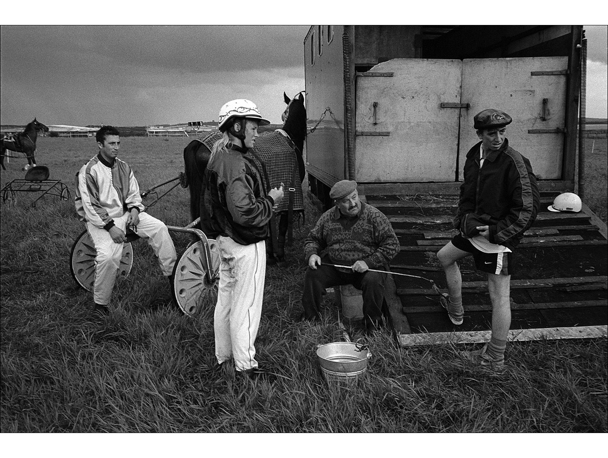 At the back of a horse box, an old man in a flat cap sits holding a whip while a young man in a Scottish cap stands next to him, a woman driver in silks stands the other side, and a male driver in silks sits on the back of a trap, as a storm gathers.