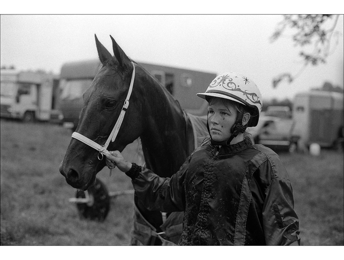 The portrait of a young blonde woman in a helmet and silk colours, stands holding the noseband of an alert horse, its ears raised, in front of a row of horse boxes, in the rain.