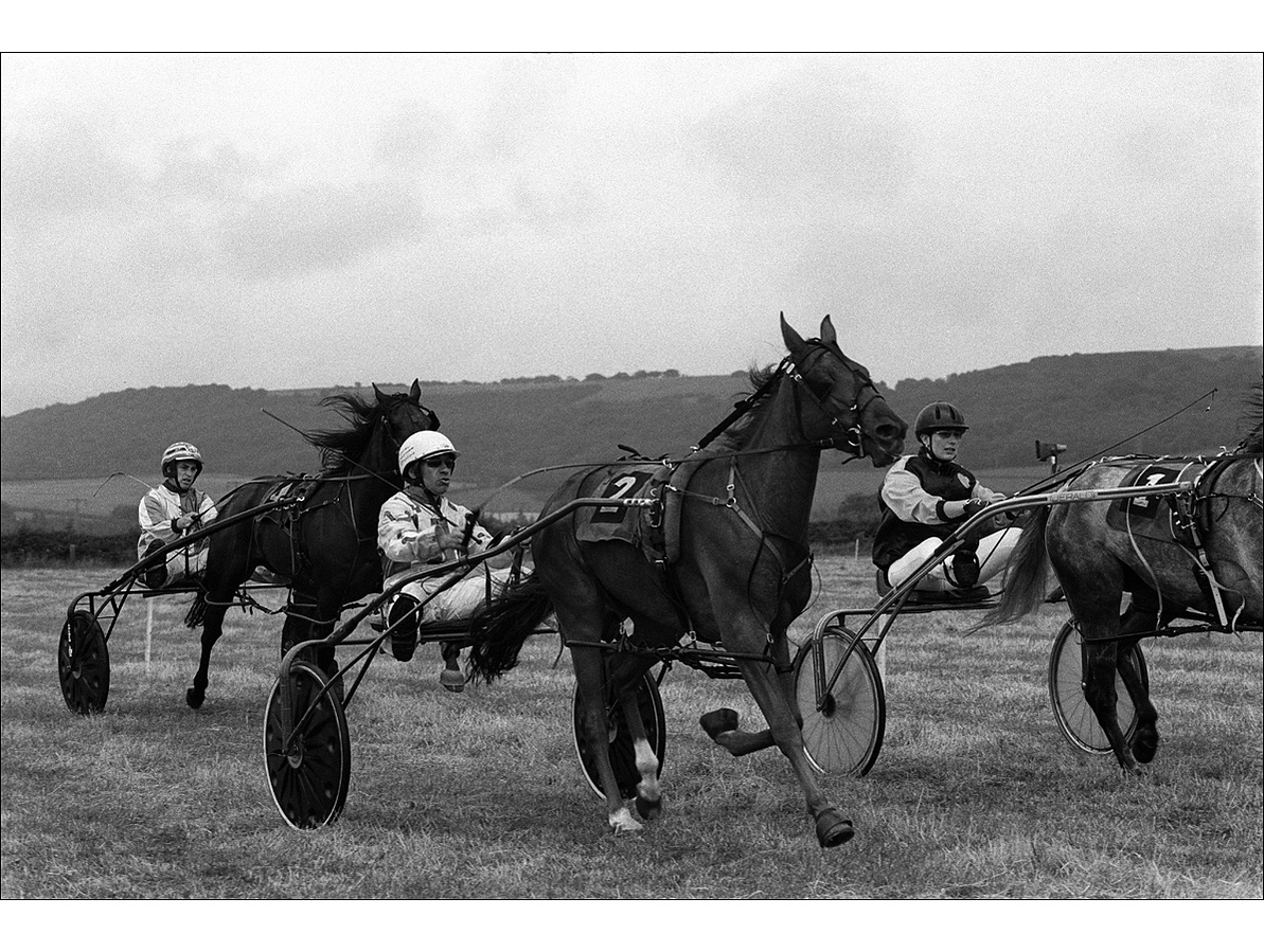 Three concentrating harness racing drivers, two men and one woman, steer their trotting horses at the start of a race in a field with tree-covered hills in the distance on a cloudy day.