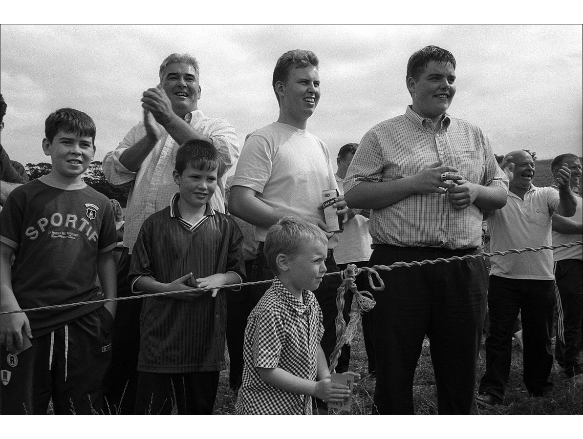 Three boys stand either side of a rope holding betting slips while five men behind them clap and cheer the harness race, in the summer sunshine.