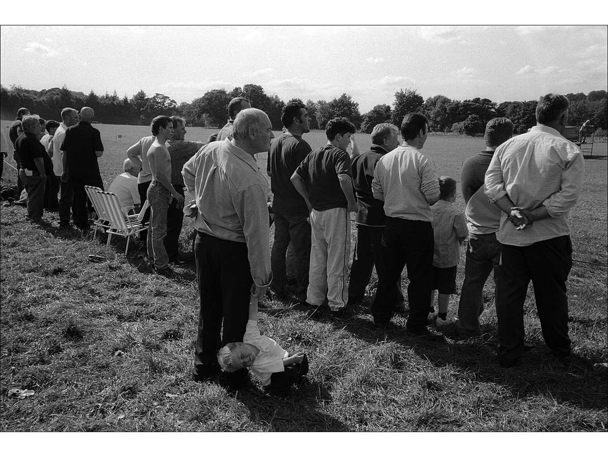 Seen from behind, a line of mostly male spectators of varying ages watch a harness race on a flat field in the countryside, while a bored little boy hangs uninterested from an older man's hand.