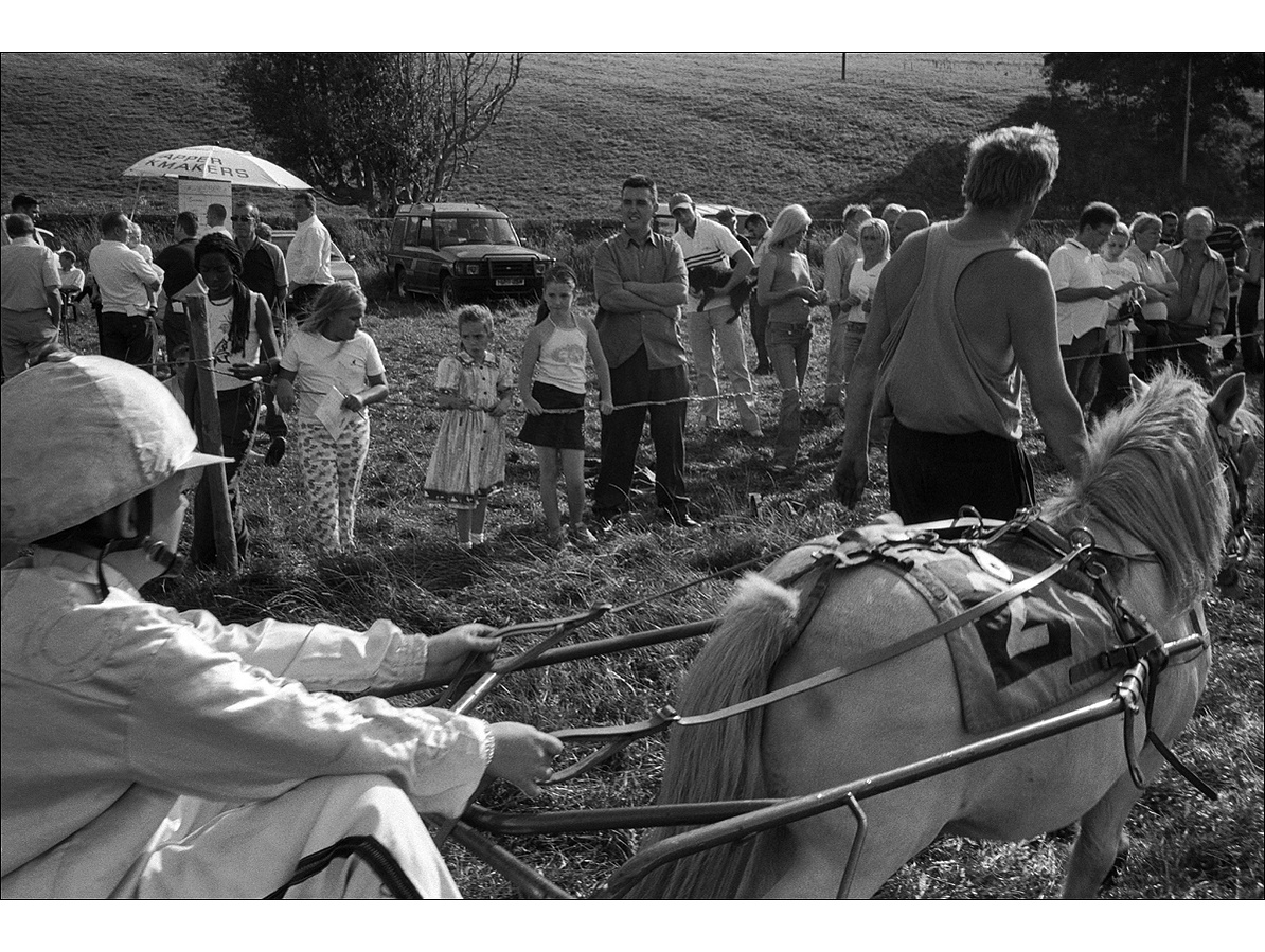 A man in a vest leads a small horse and its driver in front of spectators stood behind a rope at the side of a field, a bookmaker takes bets in the background.