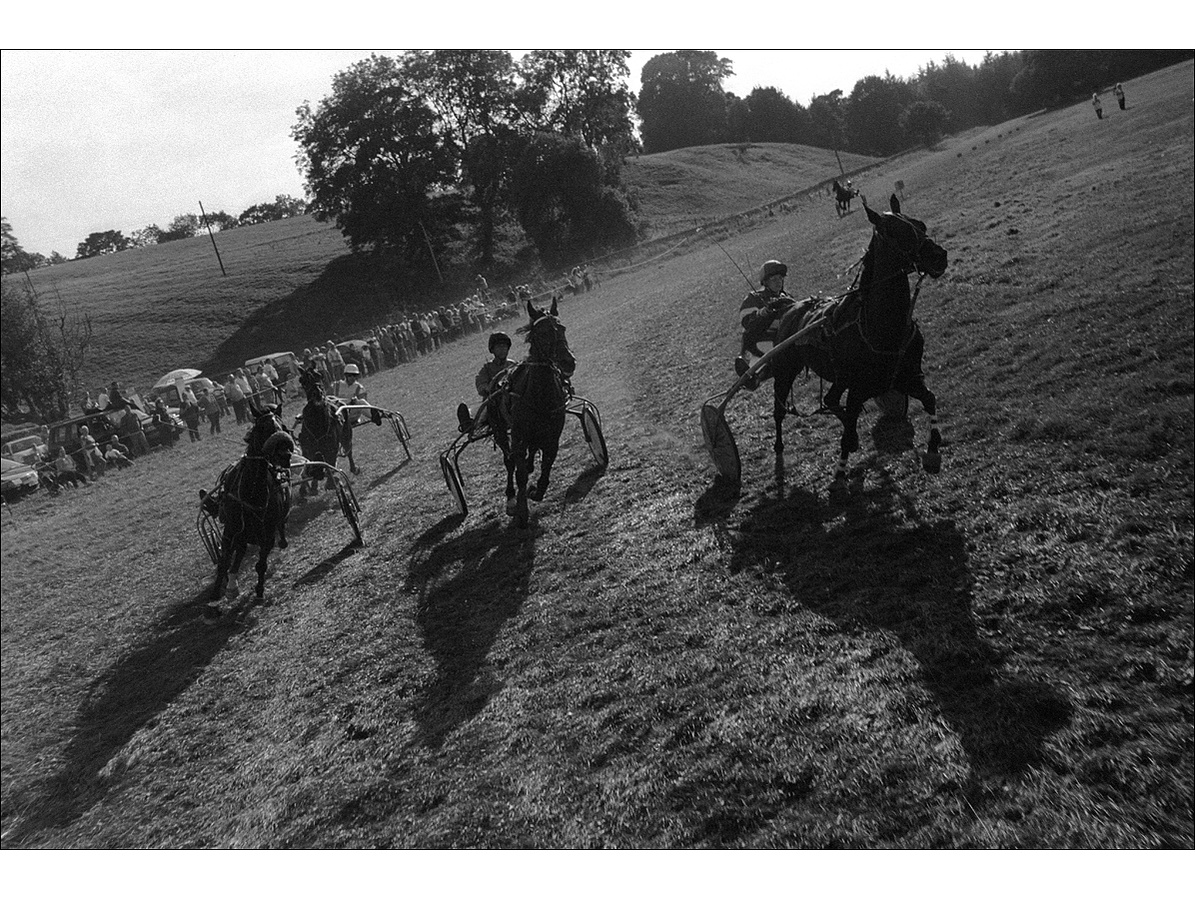 Four horses being steered around the bend of the track during a race by drivers sat on traps behind them, as spectators watch the race from the sidelines at the edge of the field.