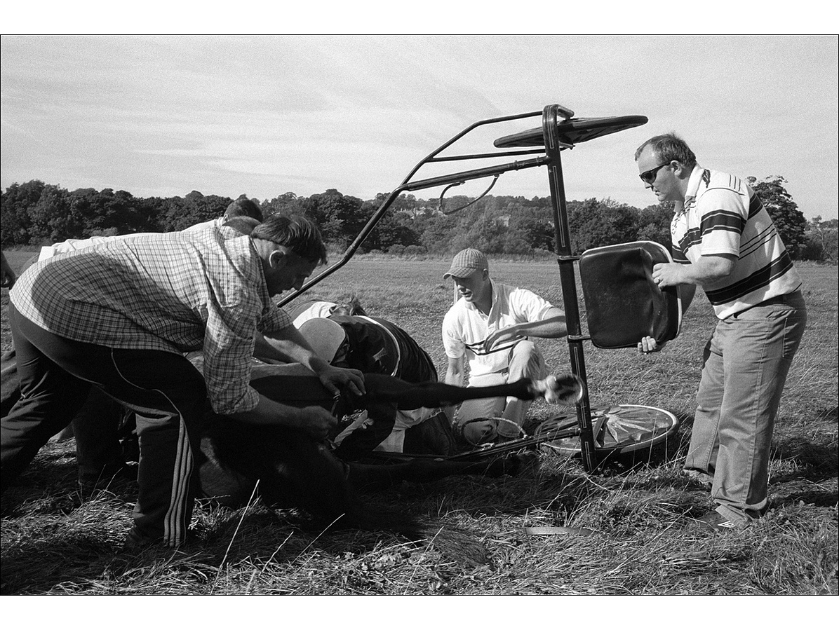 A group of concerned men gathered round a fallen and distressed horse, trying to release it from its strapping and the trap laying on its side, on a flat field in summertime.