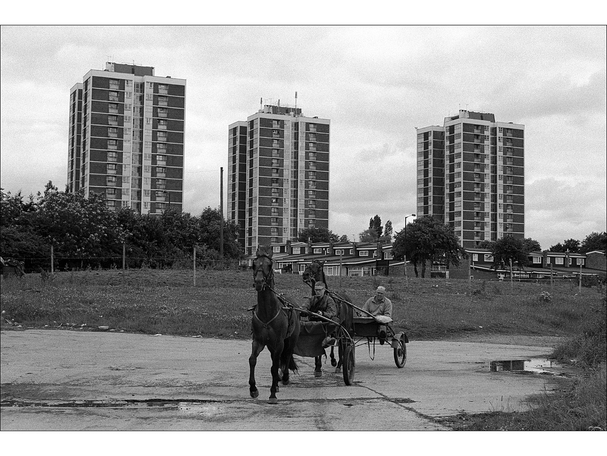 A young man drives a horse and trap followed by an older man, across a concrete area, as three tower blocks dominate the skyline behind them.