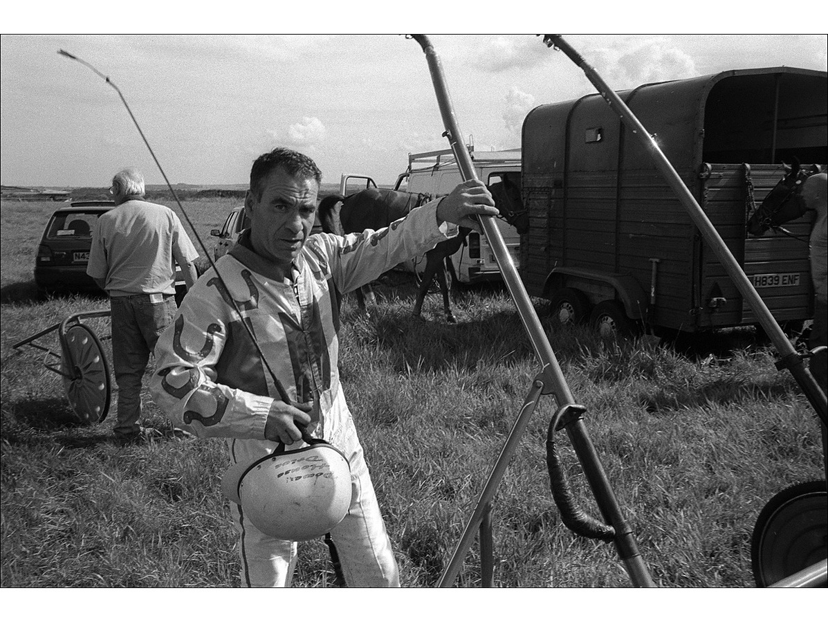 A harness driver in his silk colours hold his whip and helmet in his right hand while holding the arm of a cart in his other, as a horse stands behind in the shade of a horse box, in a field.