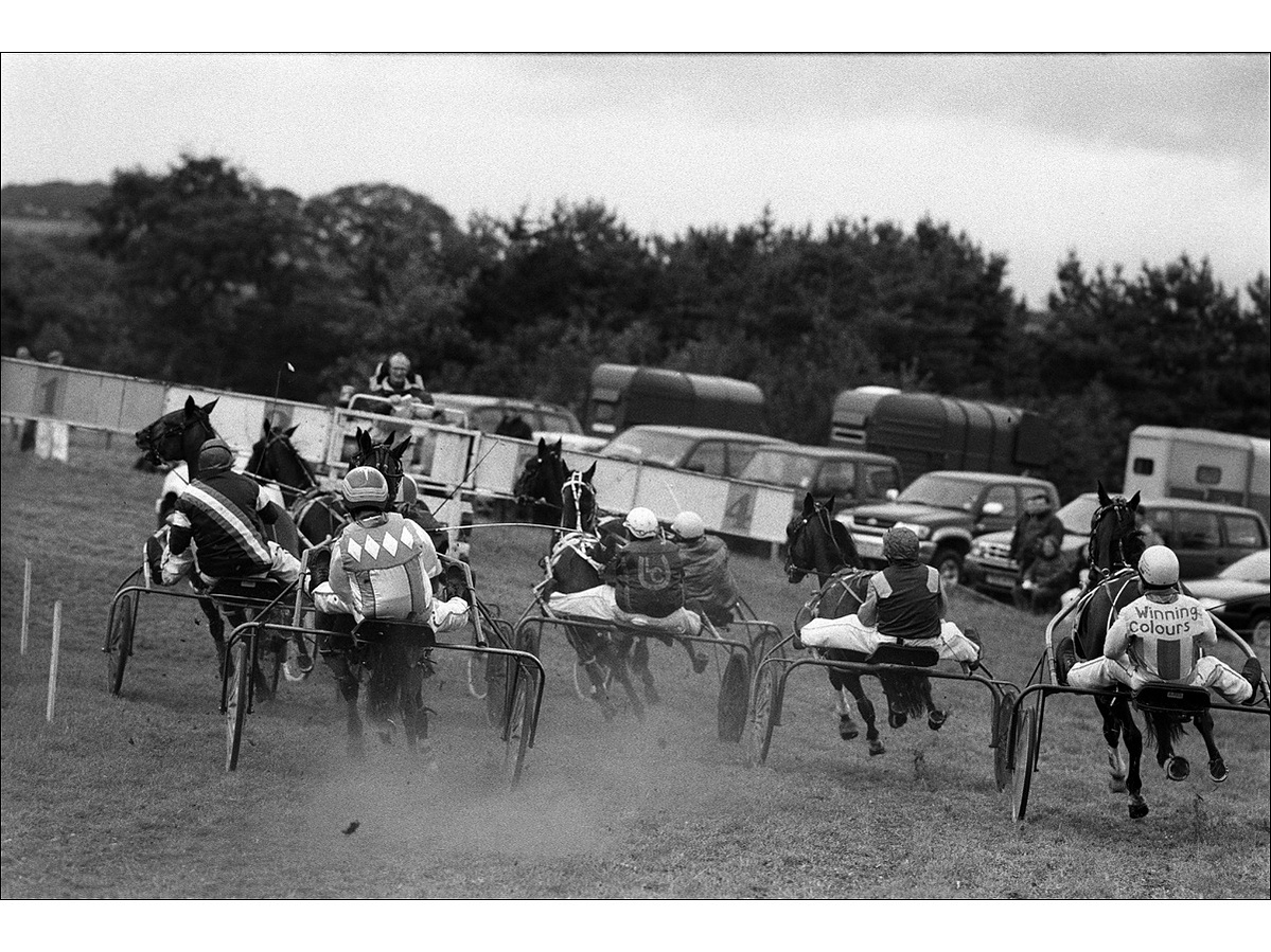 See from behind, eight harness racing horses and drivers moving at speed behind the start car, turning in the curve of the track, hovves kicking up dust and dirt, in a field.