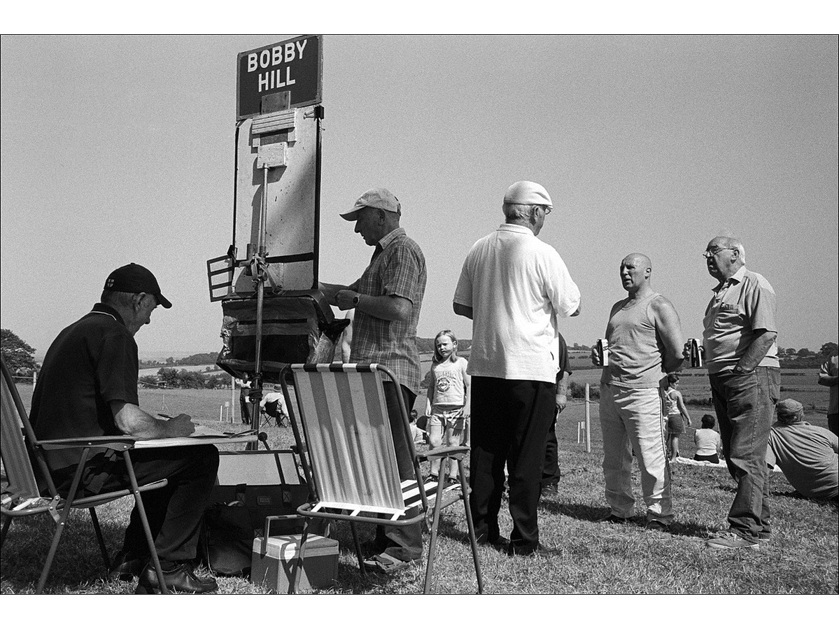 Three bookmakers setting the odds and taking bets for a harness race, for the proprietor Bobby Hill, in a rural setting in summertime, as two men stand drinking canned beer chatting with the bookmaker.