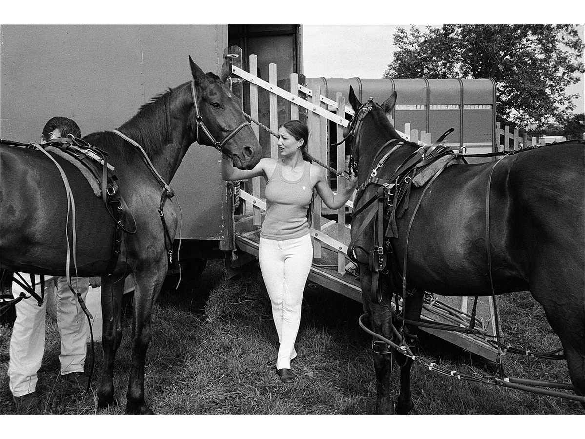 A young woman in a vest and white riding trousers holds the heads of two horses by their straps, while an older man prepares one of the horse for racing, the other is ready, in front of a horse box.