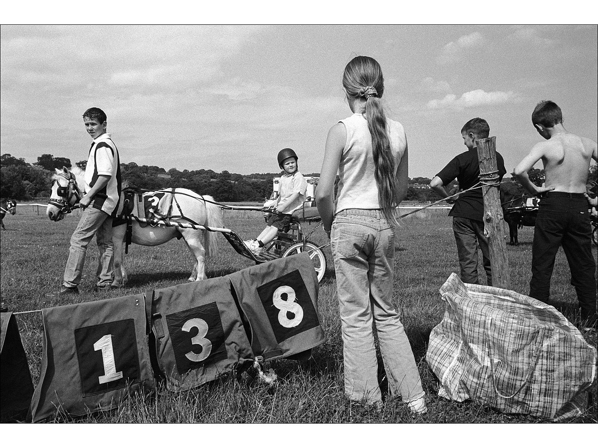 A boy sits on a trap with reins in hand, while another boy holds the white horse stationery. Three other children watch on next to a rope from which the runners numbers are hanging.