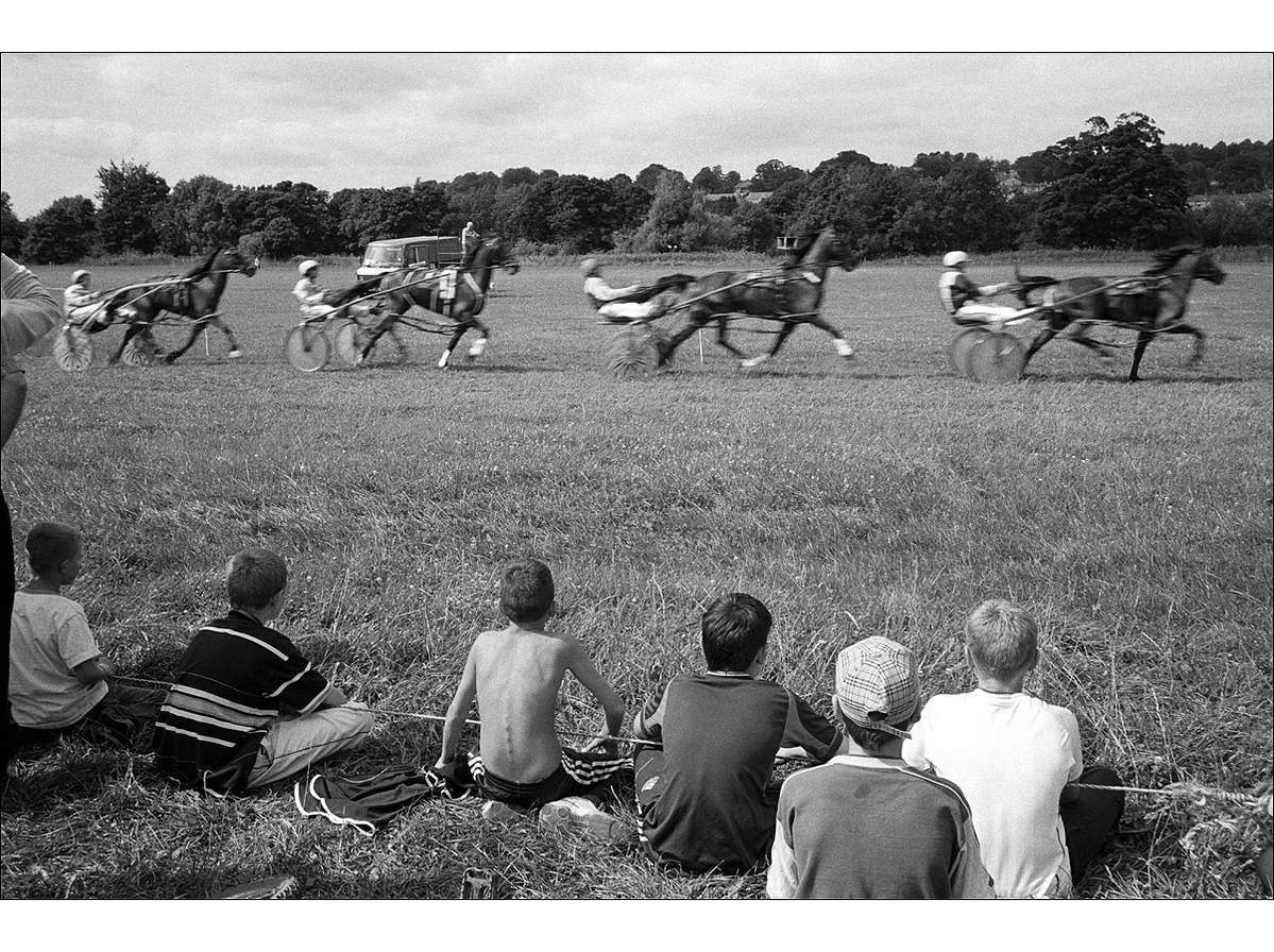 Six boys sitting on dry grass behind a rope watching four harness racing horses and drivers speeding past so quickly that they're blurred, in a flat field in summertime.