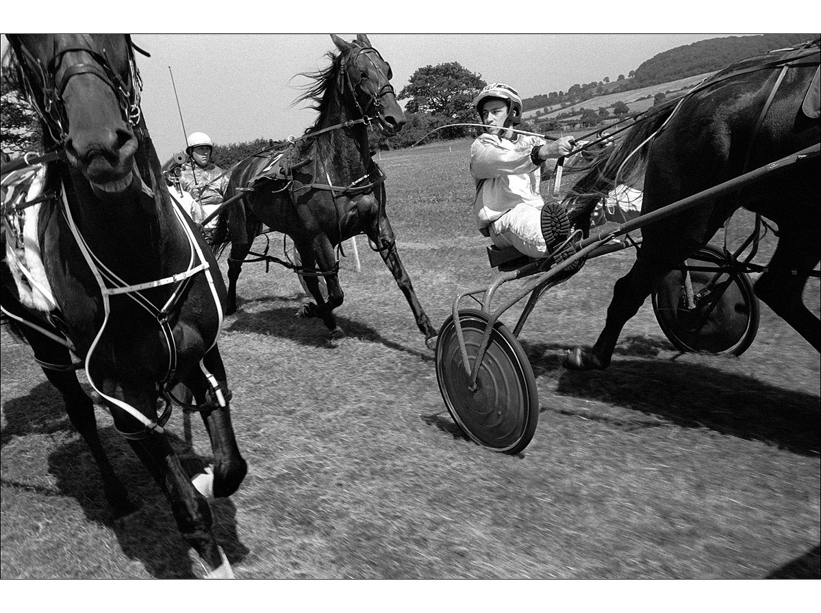 In a rural setting in summertime, two harness race drivers in colours and helmets steer their horse by their reins with whips in their right hands, while another horse and trap runs to their right.