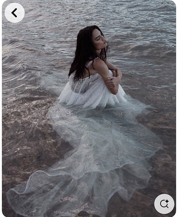 Female model crouched down at the seashore wearing a long white floating dress, hugging her knees with eyes closed 