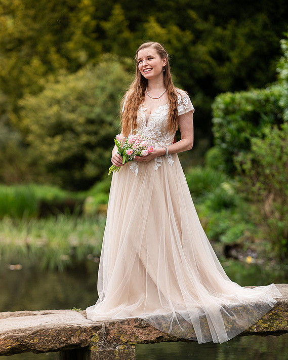 Female model Jade wearing a floor length cream coloured wedding dress, holding a small bouquet of pink flowers, standing on a stone bridge over a small river with a woodland background 