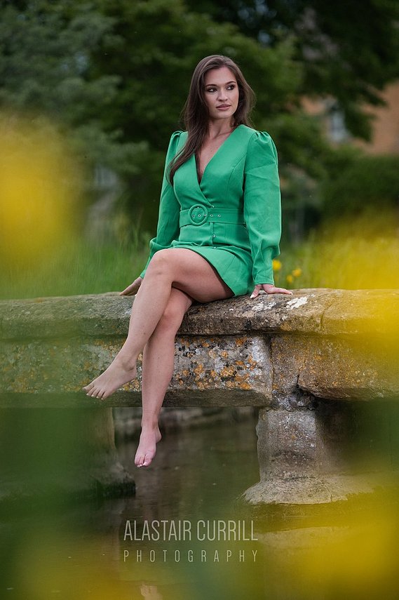 Female model Gemma Clarke wearing a green blazer sat on a stone bridge over a river in Lower Slaughter- a village in the Cotswolds