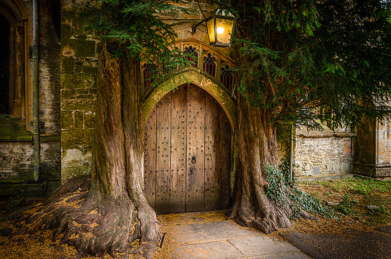 The famous wooden brown church door that inspired the Hobbit stories, ancient trees either side of the door with green leaves and a lamp handing above the door 