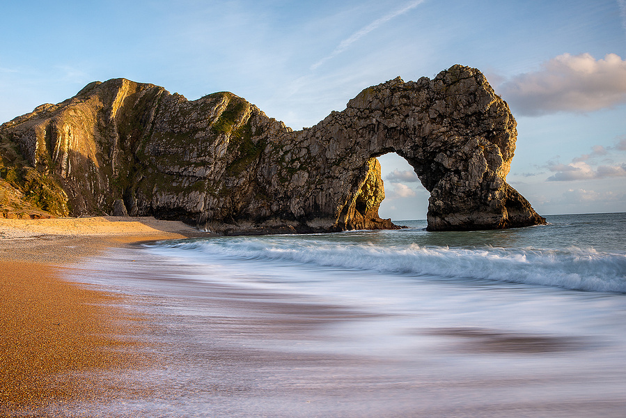 Durdle door arch with golden sand in the foreground, waves coming in and blue skies with some white cloud