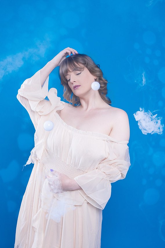 Female model wearing cream coloured floaty dress, posing with right hand on head and left hand resting over her stomach, standing against a blue backdrop with smoke machine affect 