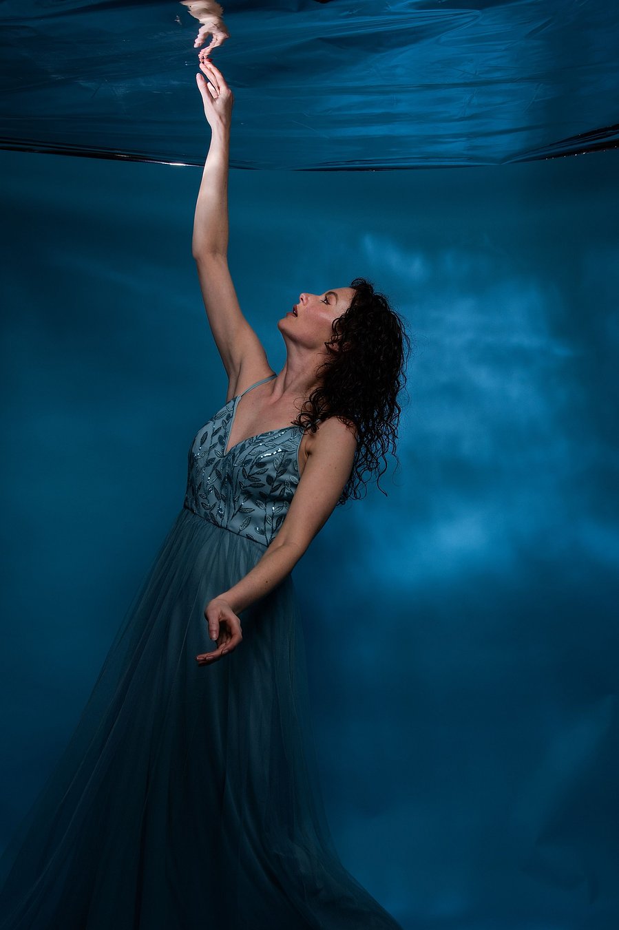 female model standing with right arm reaching upwards posed, wearing a blue floor length dress with sequin detail stood against blue shimmer backdrop