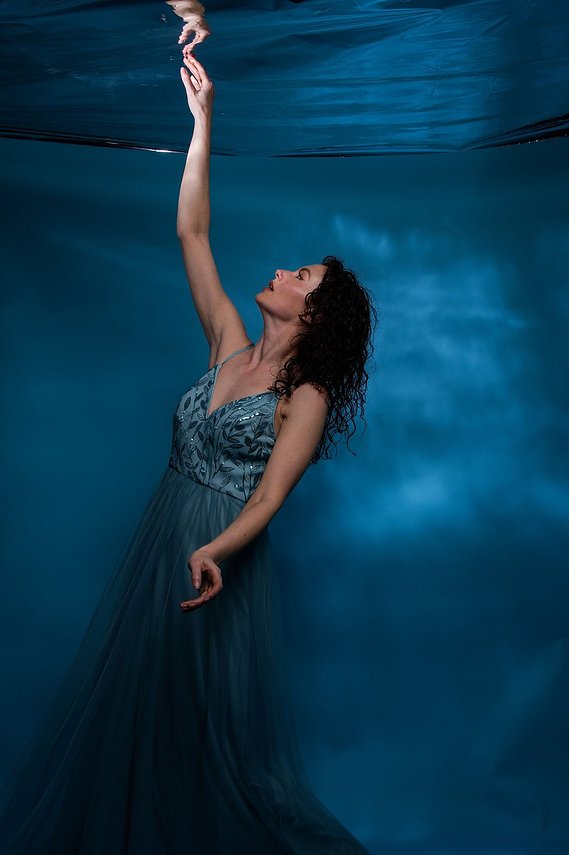 Female model wearing floor length blue dress with sequin detailing, posing with right hand reaching upwards against a blue studio backdrop 
