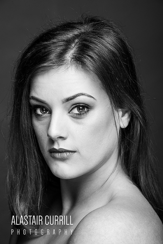 Female model headshot in black and white, looking directly at the camera with a grey background, watermark of Alastair Currill Photography along the bottom edge 
