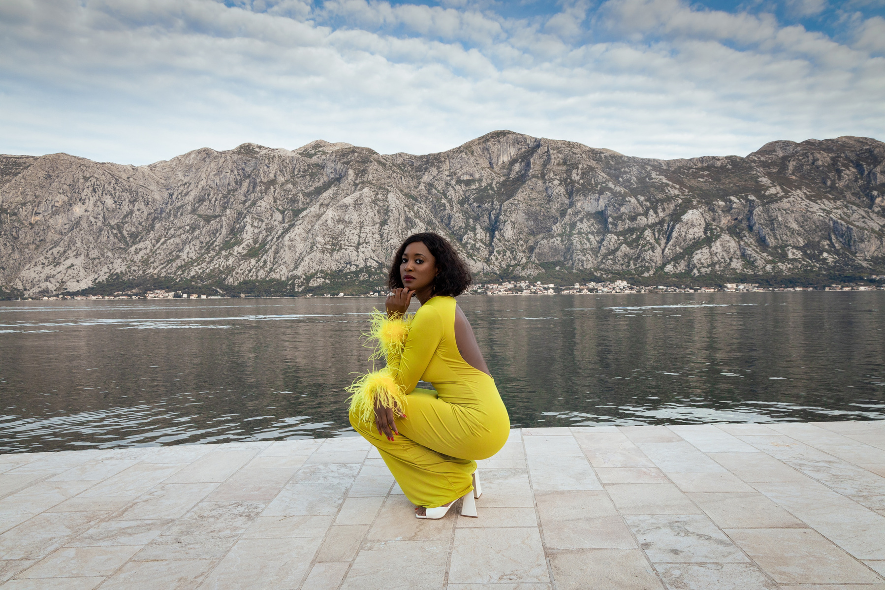 Nwando Ebeledike wearing a green dress with feather sleeves and squatting in front of a mountain by the water in Montenegro