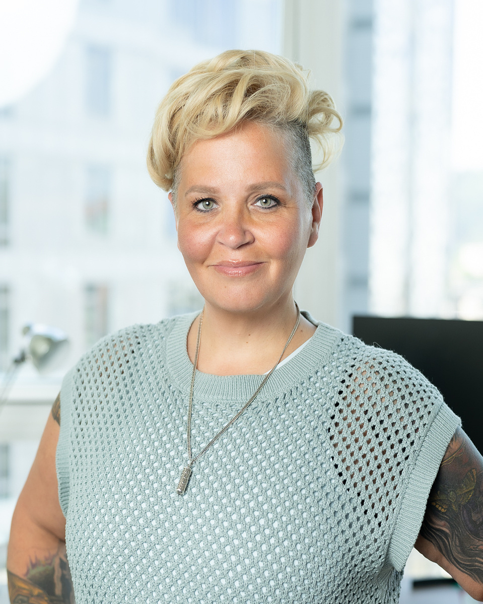 A white woman with blonde hair stands in her apartment for a headshot