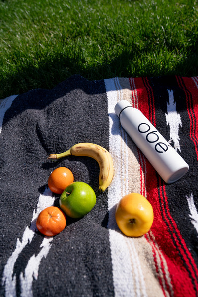 Still life photograph of fruits including banana, clementines, apples and a water bottle on a picnic blanket.