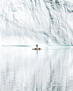 Lonesome stand up paddler in front of huge iceberg wall reflecting in the Greenland sea.