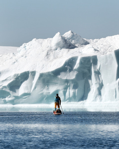 Stand up paddler in front of big iceberg wall in Greenland.